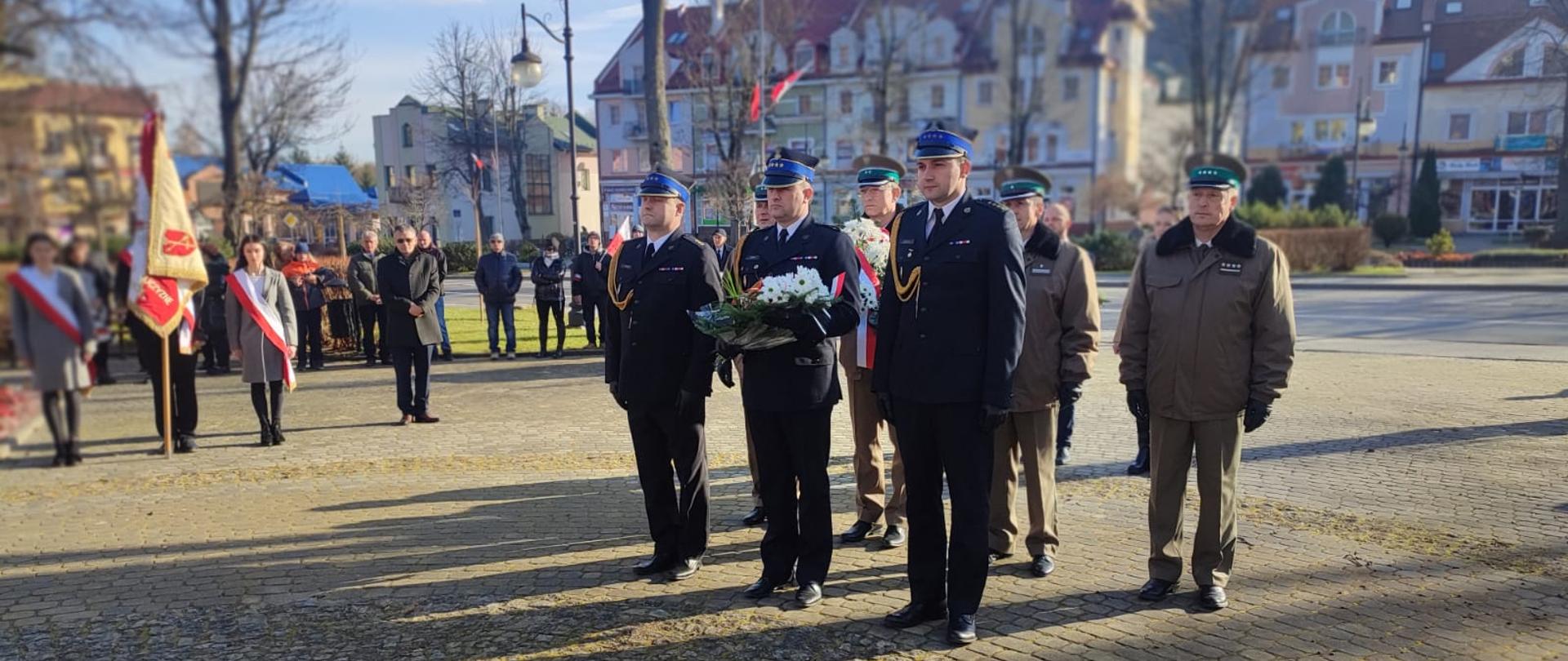 Na zdjęciu widać trzech funkcjonariuszy w mundurach galowych z KP PSP w Ustrzykach Dolnych, w tym komendanta powiatowego który stoi pomiędzy dwoma kolejnymi funkcjonariuszami. Komendant trzyma kwiaty w rękach. Za nimi stoi trzech przedstawicieli Straży Granicznej. Po lewej stronie widać poczet sztandarowy i grupę osób. W tle zabudowa miasta. Wydarzenie odbywa się na rynku w Ustrzykach Dolnych.