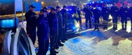 Policjanci i strażacy stoją przed swoimi samochodami służbowymi, część z nich salutuje. W tle lampy oświetlenia ulicznego, znaki drogowe. Na ziemi leży śnie, aura nocna.