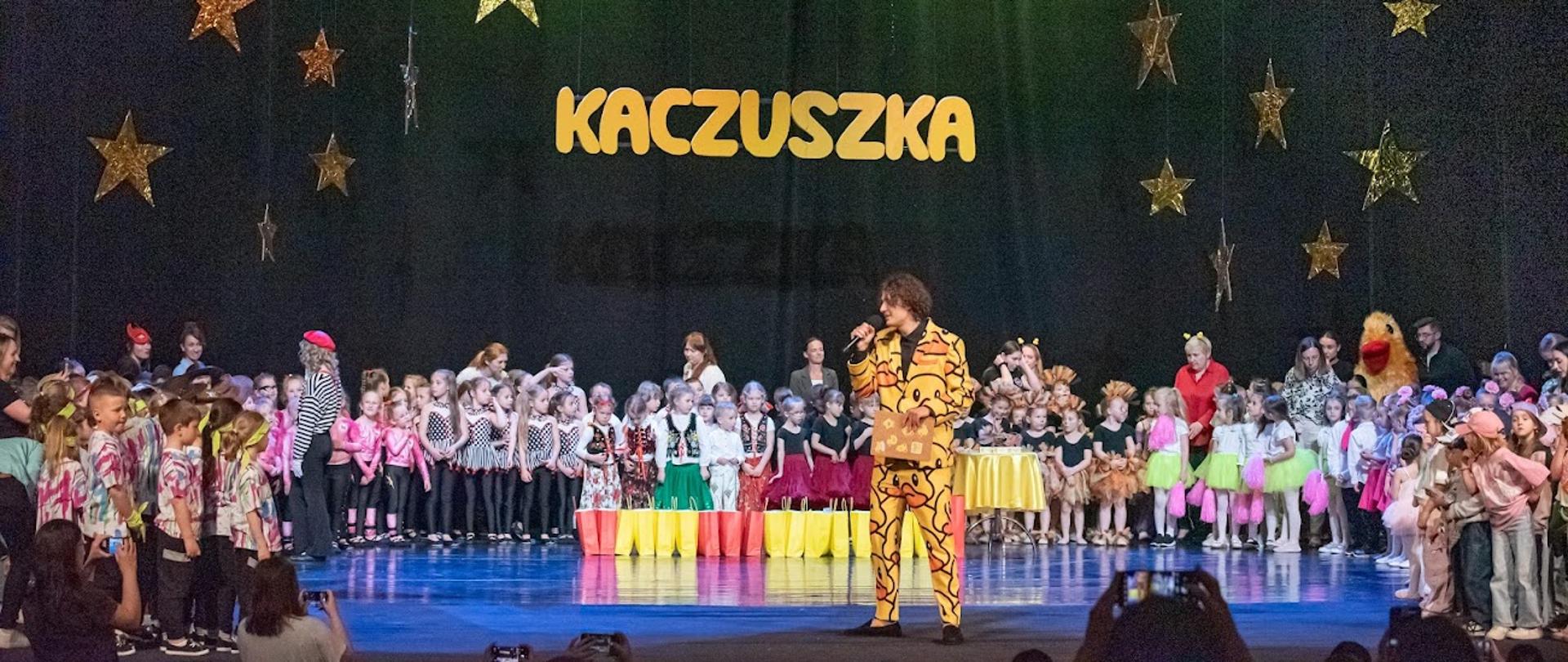Zdjęcie przedstawia dużą grupę dzieci oraz ich opiekunów na scenie. Na pierwszym planie stoi słuchacz Studium, ubrany w żółto-czarny garnitur oraz trzyma mikrofon.