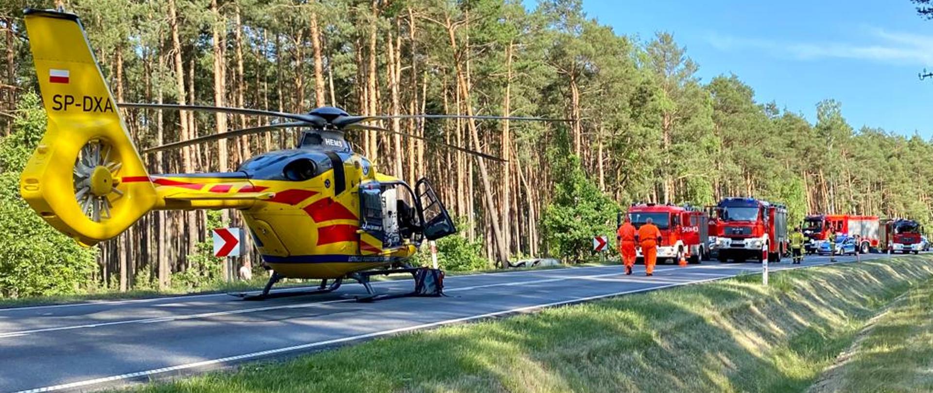 Zdjęcie przedstawia akcję ratowniczą wypadku drogowego z udziałem samochodu osobowego na drodze krajowej nr 11. Na zdjęciu widać śmigłowiec lotniczego pogotowia ratunkowego oraz wozy ratowniczo - gaśnicze. Zdjęcie wykonane w porze dziennej na poboczu drogi krajowej.