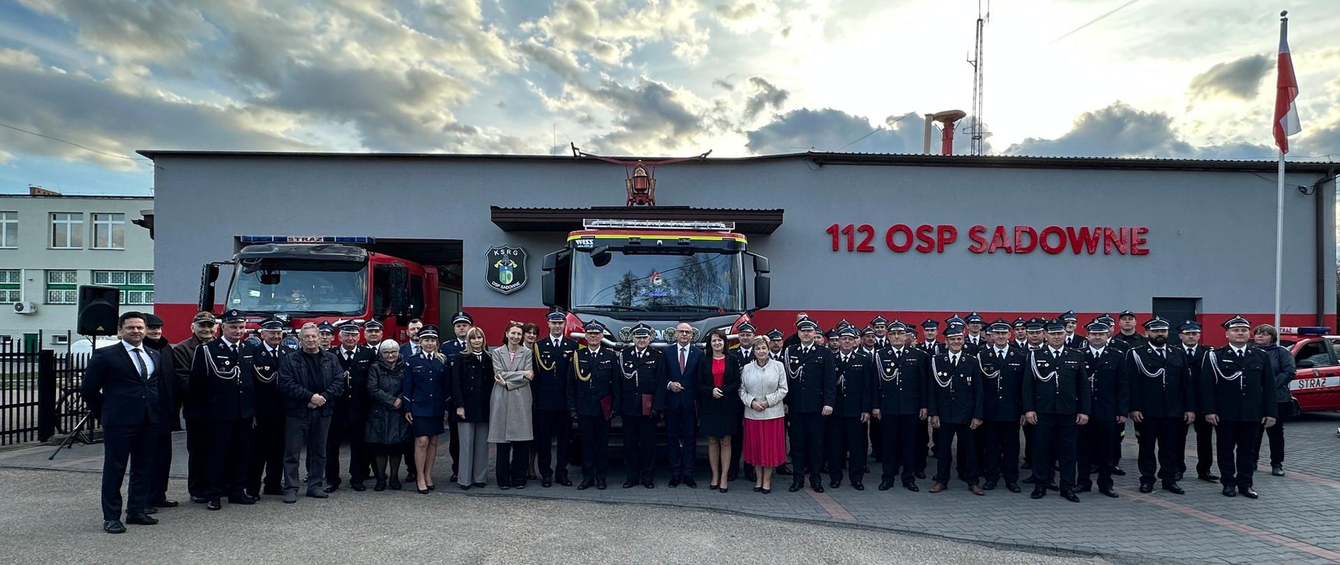 Zdjęcie grupowe uczestników uroczystości stojących na tle samochodów strażackich i remizy na której widniej napis "112 OSP Sadowne" 