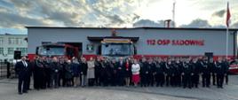 Zdjęcie grupowe uczestników uroczystości stojących na tle samochodów strażackich i remizy na której widniej napis "112 OSP Sadowne" 
