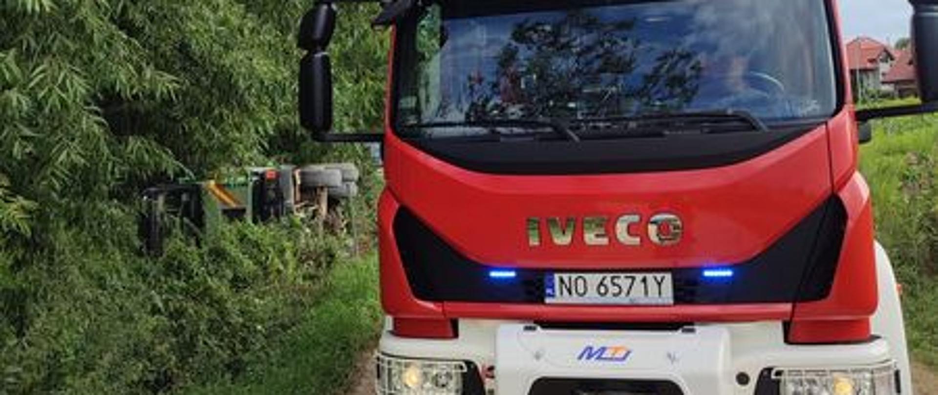 Samochód gaśniczy stoi na drodze polnej za nim widać samochód ciężarowy który znajduje się na boku w rowie. 