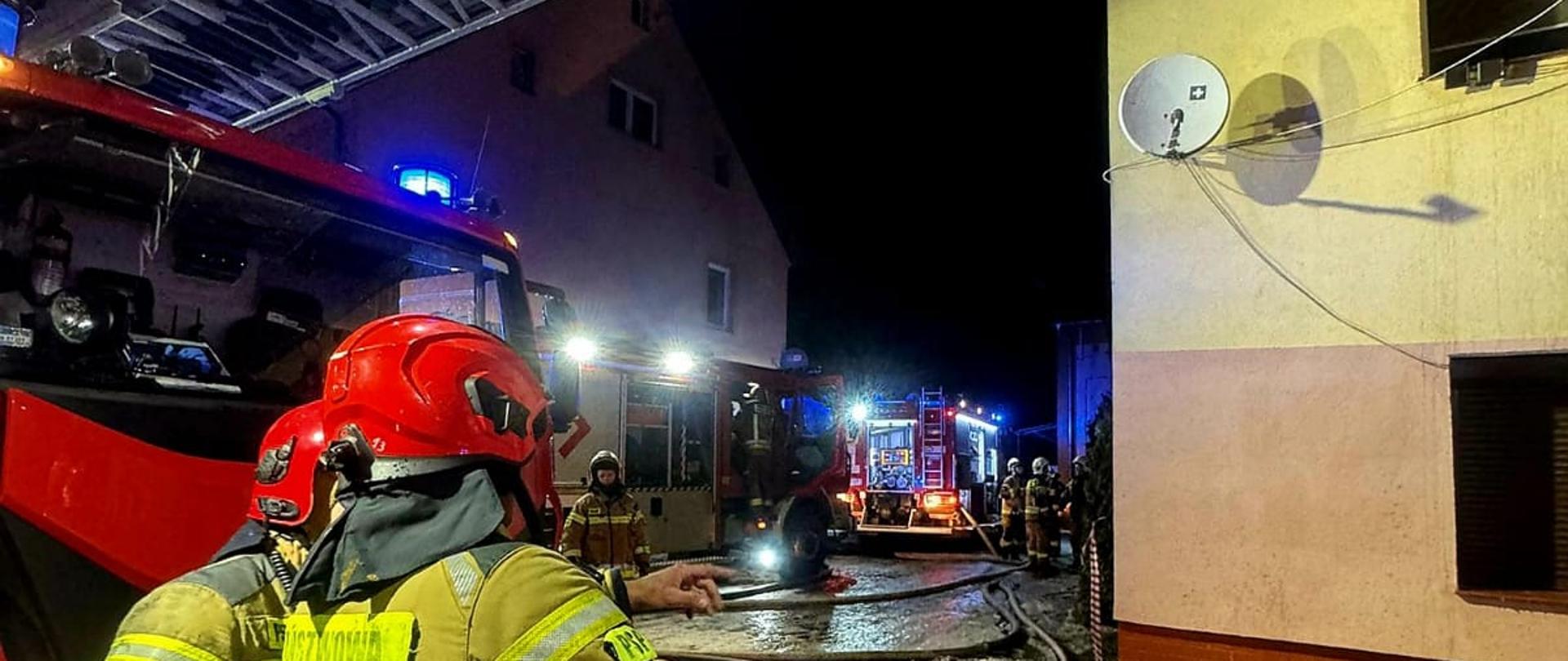 Zdjęcie przedstawia scenerię w trakcie lub pod koniec akcji gaśniczej prowadzonej w warunkach nocnych na terenie zabudowanym.
Oto szczegółowy opis elementów widocznych na zdjęciu:
Strażacy na pierwszym planie: Po lewej stronie kadru widoczne są sylwetki dwóch strażaków Państwowej Straży Pożarnej (PSP) w pełnym umundurowaniu bojowym (piaskowo-musztardowe ubrania specjalne z elementami odblaskowymi) oraz czerwonych kaskach. Strażak bliżej obiektywu jest obrócony plecami i wskazuje ręką w kierunku strefy działań, natomiast drugi stoi tuż obok niego.
Sprzęt i pojazdy: * W górnej części kadru, nad głowami strażaków i nad drogą, rozciąga się metalowa konstrukcja wysuniętej drabiny mechanicznej.
Po lewej stronie widać fragment czerwonego wozu strażackiego, a w głębi ulicy zaparkowane są kolejne pojazdy ratownicze z włączonymi, niebieskimi sygnałami błyskowymi.
Otoczenie i skutki pożaru:
Po prawej stronie stoi piętrowy budynek mieszkalny z jasną elewacją i zamontowaną anteną satelitarną.
Na ziemi (chodniku i drodze) leży mnóstwo potłuczonych, czerwonych dachówek ceramicznych, które spadły z płonącego dachu, a także resztki stopionego śniegu i lodu.
Przez całą szerokość drogi i wzdłuż budynków rozciągnięte są liczne, białe węże strażackie (linie gaśnicze i zasilające). Teren bezpośrednio pod ścianą domu został odgrodzony biało-czerwoną taśmą ostrzegawczą.
Oświetlenie: Scena jest dynamicznie oświetlona mieszanką zimnego światła z reflektorów wozów strażackich oraz intensywnych, niebieskich błysków kogutów, co nadaje ujęciu reporterski charakter.