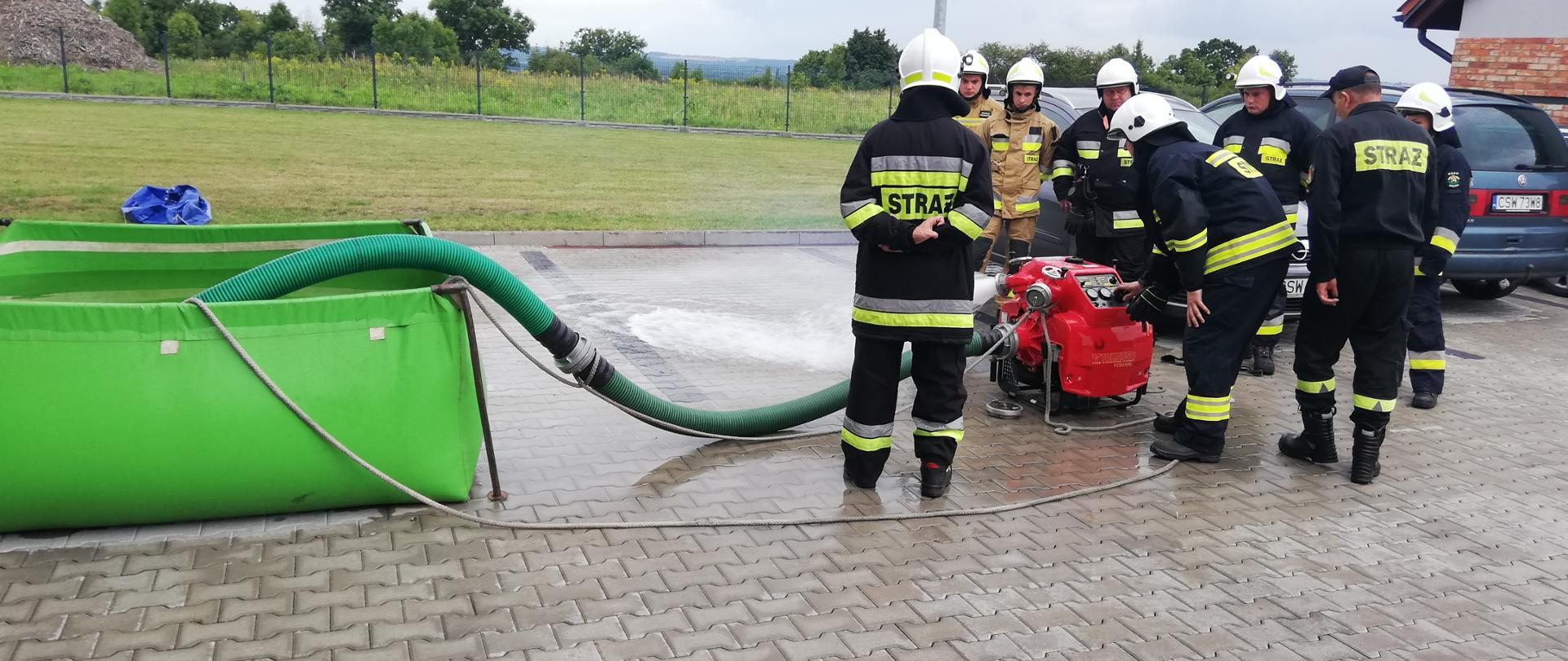 Po prawej stronie zdjęcia wokół motopompy pożarniczej stoją strażacy. Jeden z nich obsługuje motopompę i przy pomocy węży ssanych zasysa wodę ze zbiornika który stoi po lewej stronie. 