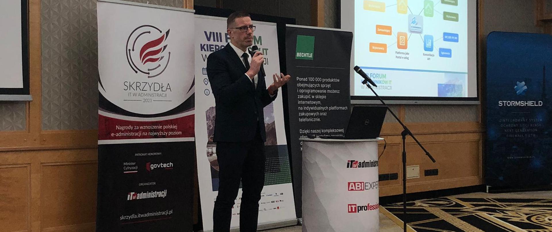 VIII Forum Kierowników IT w Administracji, Krzysztof Fijołek, Dyrektor Generalny UZP przemawia do uczestników wydarzenia trzymając w ręku mikrofon. W tle pokazany jest slajd prezentacji.