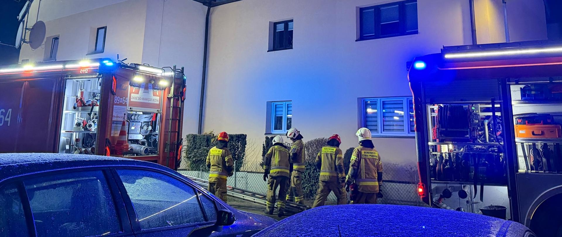 Nocna akcja straży pożarnej przed jasnym budynkiem mieszkalnym; kilku strażaków w hełmach stoi przy wozach strażackich z otwartymi skrytkami, migają niebieskie światła, na pierwszym planie zaparkowane samochody.