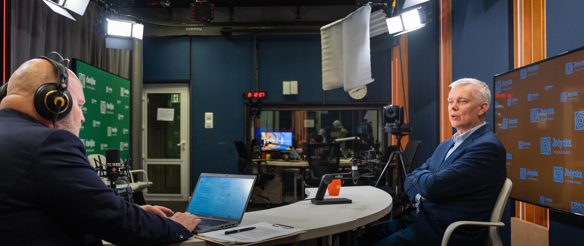 Wnętrze studia radiowego. Przy okrągłym stole siedzą dwaj mężczyźni podczas rozmowy. Po lewej stronie znajduje się prowadzący w słuchawkach, pracujący przy laptopie i mikrofonie. Po prawej siedzi minister Tomasz Siemoniak w niebieskiej marynarce, również przy mikrofonie. Wokół widoczne są lampy studyjne, kamery, sprzęt nagraniowy oraz monitory z logo Jedynki Polskiego Radia.