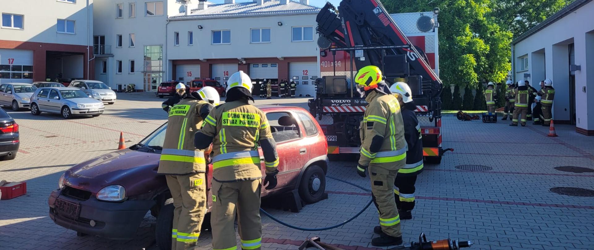 Grupa 4 strażaków z Ochotniczej Straży Pożarnej ubranych w umundurowanie specjalne koloru piaskowego używając specjalistycznego sprzętu hydraulicznego próbuje wywarzyć przednie prawe drzwi do samochodu osobowego koloru czerwonego. Dwóch strażaków Komendy Powiatowej PSP w Bochni nadzoruje zadane ćwiczenie w ramach egzaminu. W pobliżu znajduje się również duży samochód strażacki. Całe ćwiczenie odbywa się na placu wewnętrznym Komendy PSP w Bochni