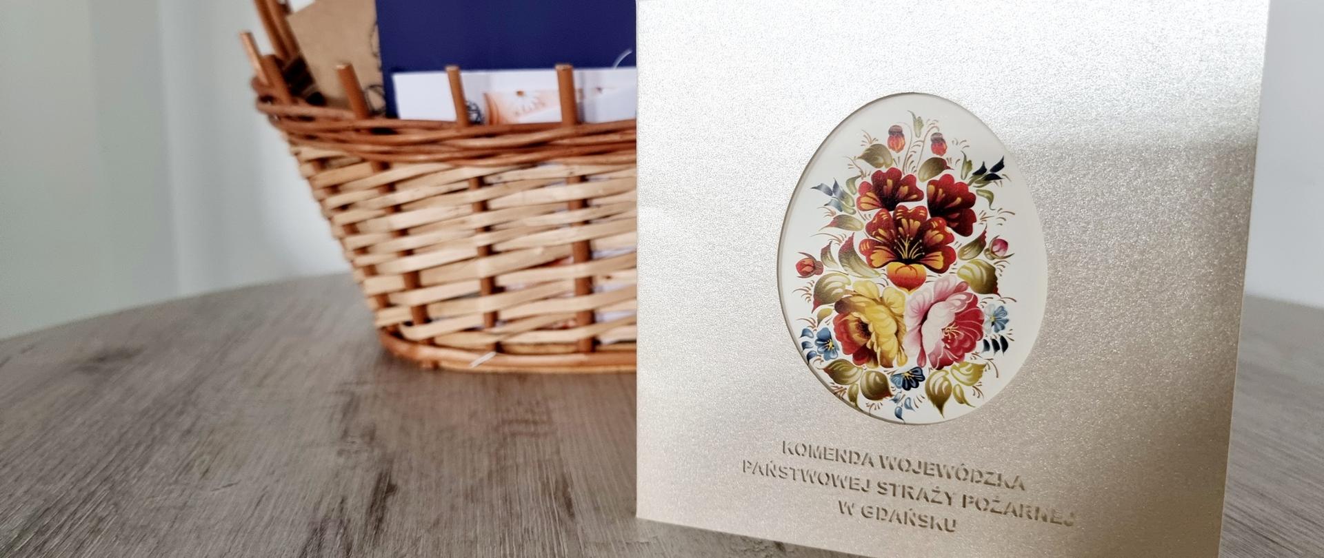 Na stoliku położony jest kosz z kartkami z życzeniami przed koszem stoi kartka z życzeniami kierownictwa Komendy Wojewódzkiej PSP.
