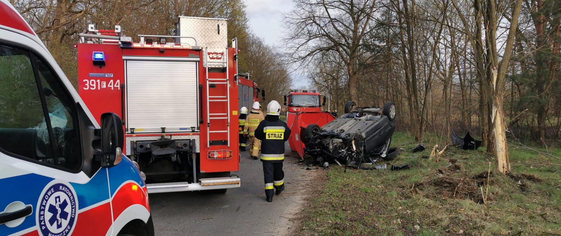 Widok leżącego na dachu uszkodzonego samochodu osobowego. Po lewej stronie stojące samochody pożarnicze oraz karetka pogotowia. Za pojazdem rozstawiony czerwony parawan. Z prawej strony na trawie widoczne rozrzucone elementy pojazdu. Na jezdni obok pojazdu umundurowani strażacy.