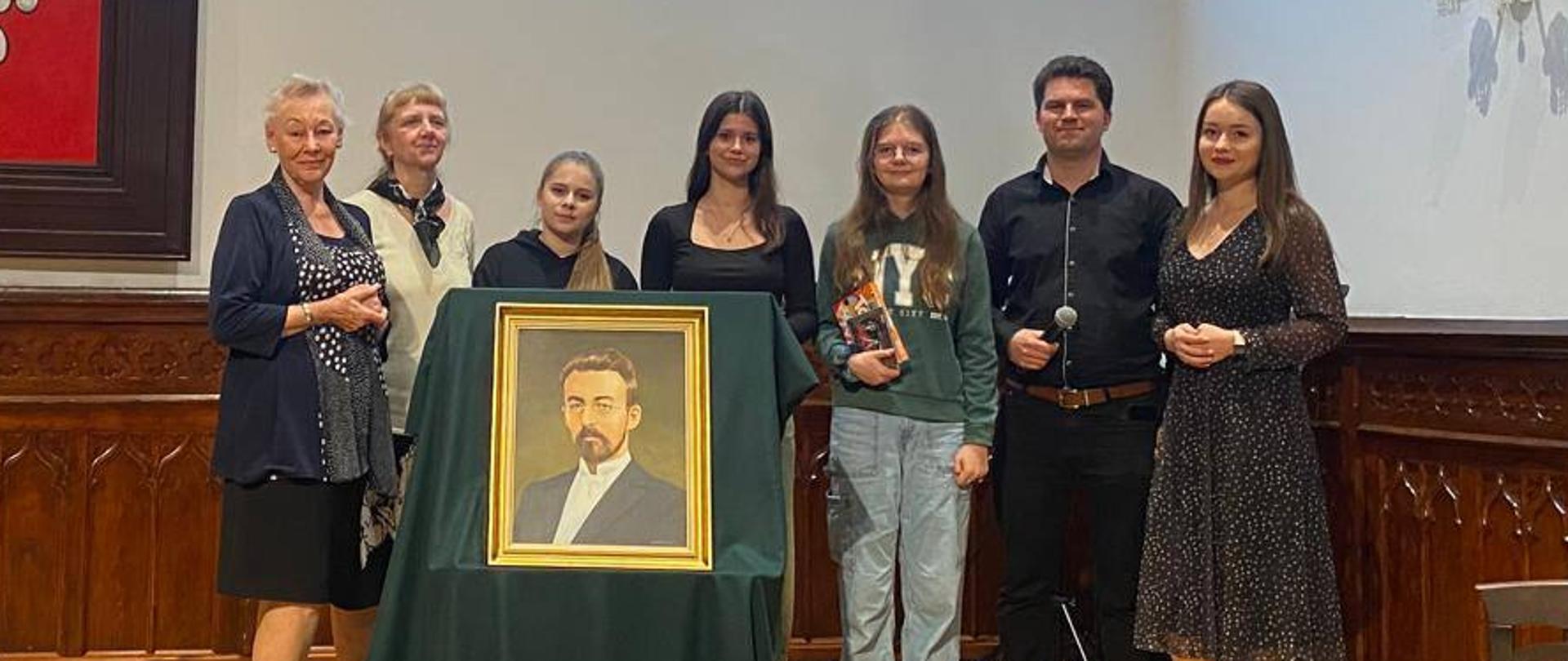 prelegenci przy portrecie Mieczysława Karłowicza na scenie w sali królewskiej PSM Mielec podczas spotkania poświęconemu patronowi mieleckiej szkoły muzycznej – Mieczysławowi Karłowiczowi