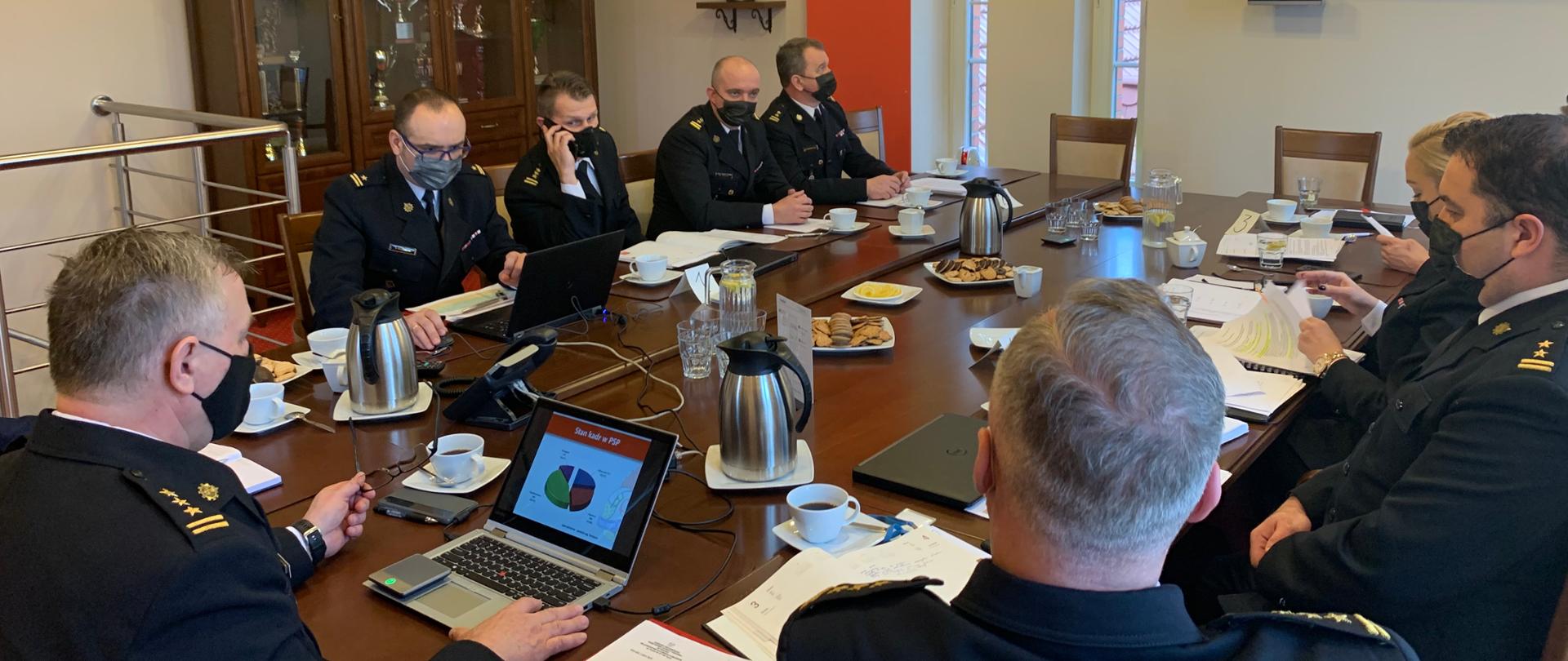 Na zdjęciu widoczni są strażacy w umundurowaniu wyjściowym, siedzący wokół stołu konferencyjnego. Wśród nich wojewoda kujawsko-pomorski. Na stole leżą notatniki oraz sprzęt techniczny, Umożliwiający prezentowanie obrazu na ekranie oraz zdalną komunikację z uczestnikami narady w powiatach. Oprócz tego na stole widać szklanki, filiżanki, napoje i ciastka. 