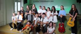 Na zdjęciu są dzieci w odświętnych strojach trzymające w rękach różne i instrumenty muzyczne. Z prawej strony stoją dwie dziewczynki trzymające w rękach gitary jest również dziewczynka i chłopiec z gitarami którzy przed nimi siedzą. Za siedzącymi klęczą czterej chłopcy którzy trzymają w rękach saksofony w kolorze złotym pomiędzy nimi siedzi dziewczynka z akordeonem. Z tyłu stoją dziewczynki trzymające w rękach skrzypce flet klarnet trąbkę. W tle na ścianie jest fragment logo szkoły