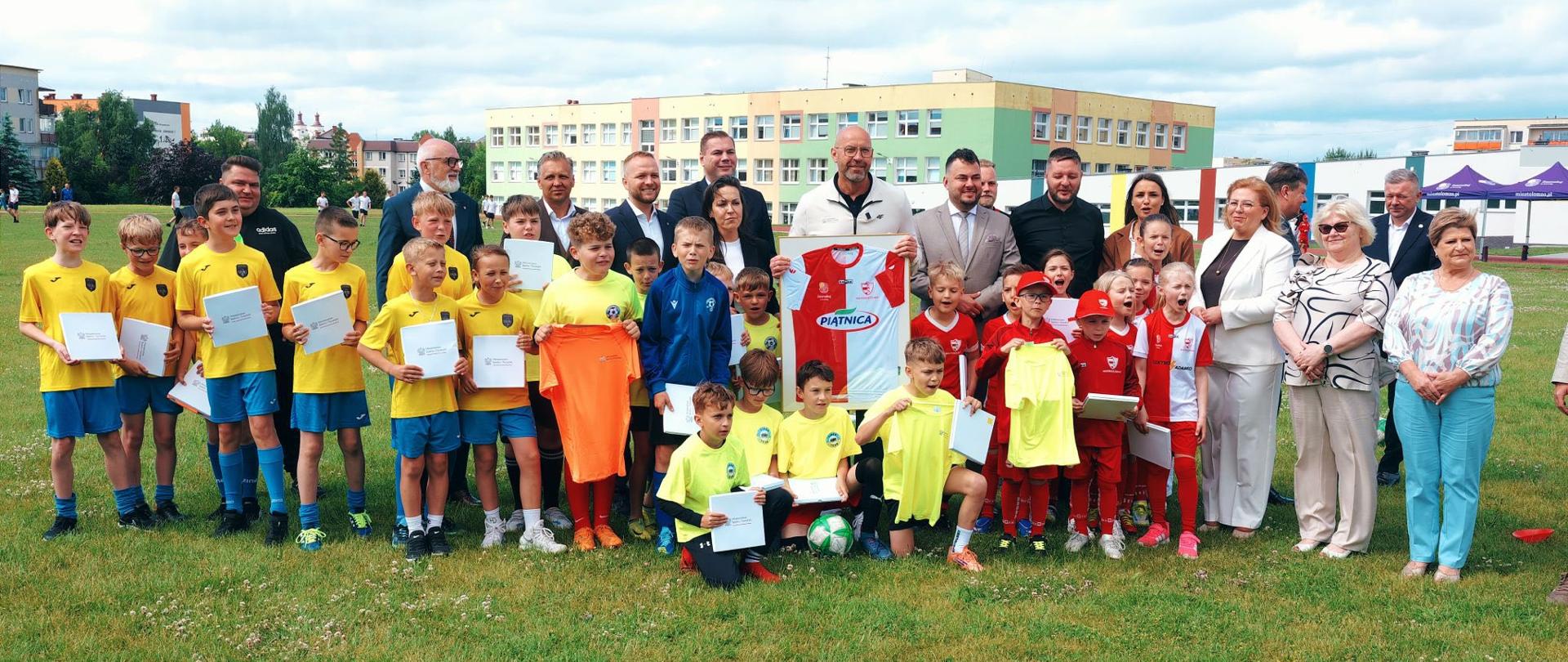 Zdjęcie grupowe na trawniku przedstawiające ministra sportu i turystyki Jakuba Rutnickiego z grupą młodych piłkarzy i piłkarek oraz przedstawicielami lokalnych władz. W tle widać budynek szkoły.