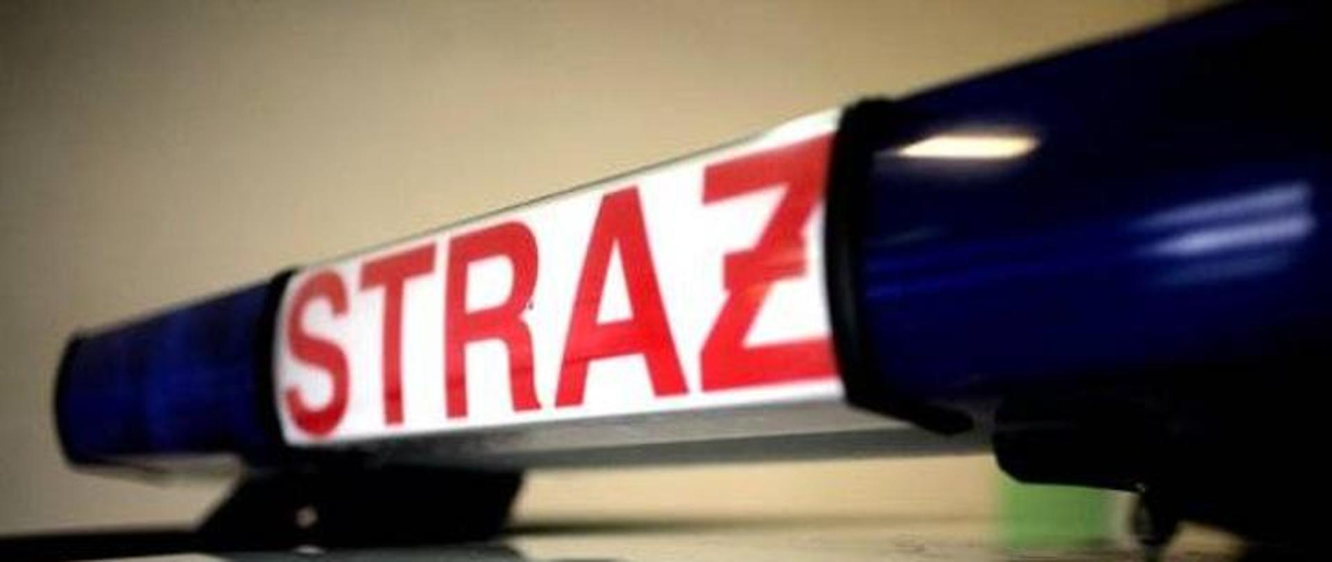 Na beżowym tle belka sygnalizacyjna z napisem straż na białym tle. Po bokach belki dwa niebieskie koguty.