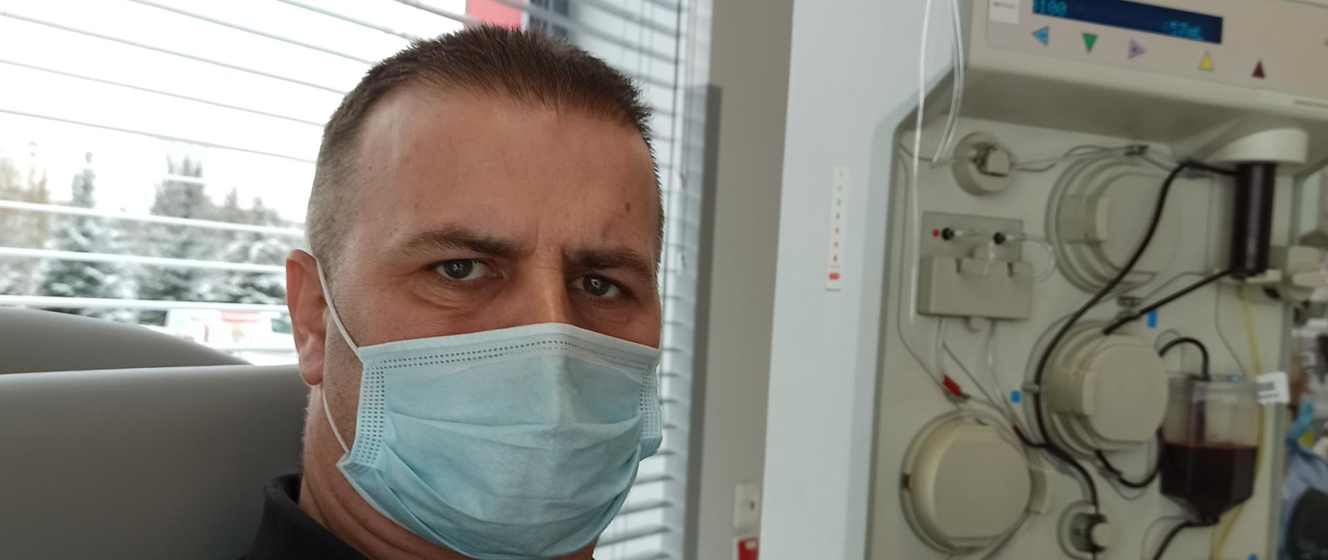 Widać strażaka w koszulce. Strażaka na lewej ręce ma założoną opaskę do mierzenia ciśnienia oraz wenflon. w tle widać aparaturę medyczną.