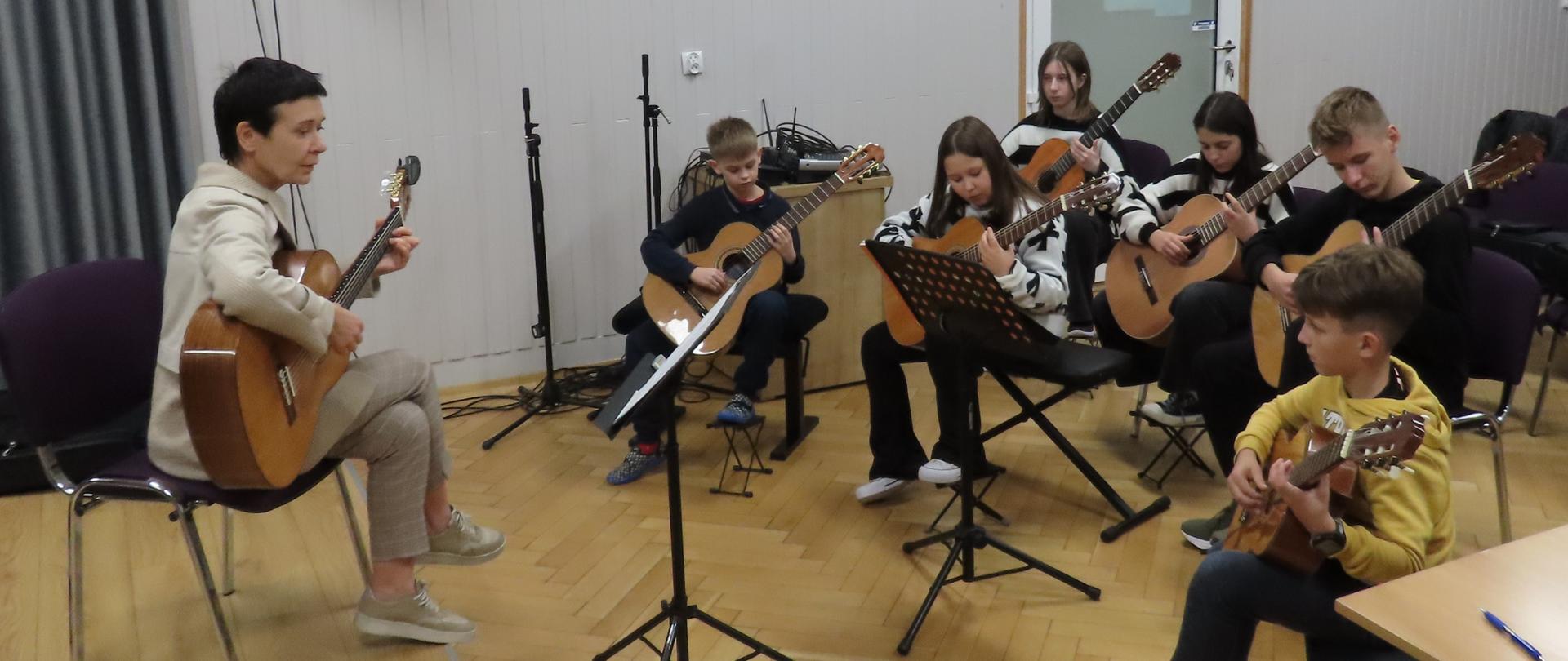 Po prawej stronie zdjęcia sześcioro dzieci w różnokolorowych ubraniach grających na gitarach. Po lewej stronie kobieta z jasnym ubraniu siedząca z nogą założoną na nogę i trzymająca w ręku gitarę. Nad jej głową zawieszona czarna kolumna głośnikowa, a obok czarno-biały portret grającego na fortepianie Witolda Lutosławskiego na tle jasnej ściany. 