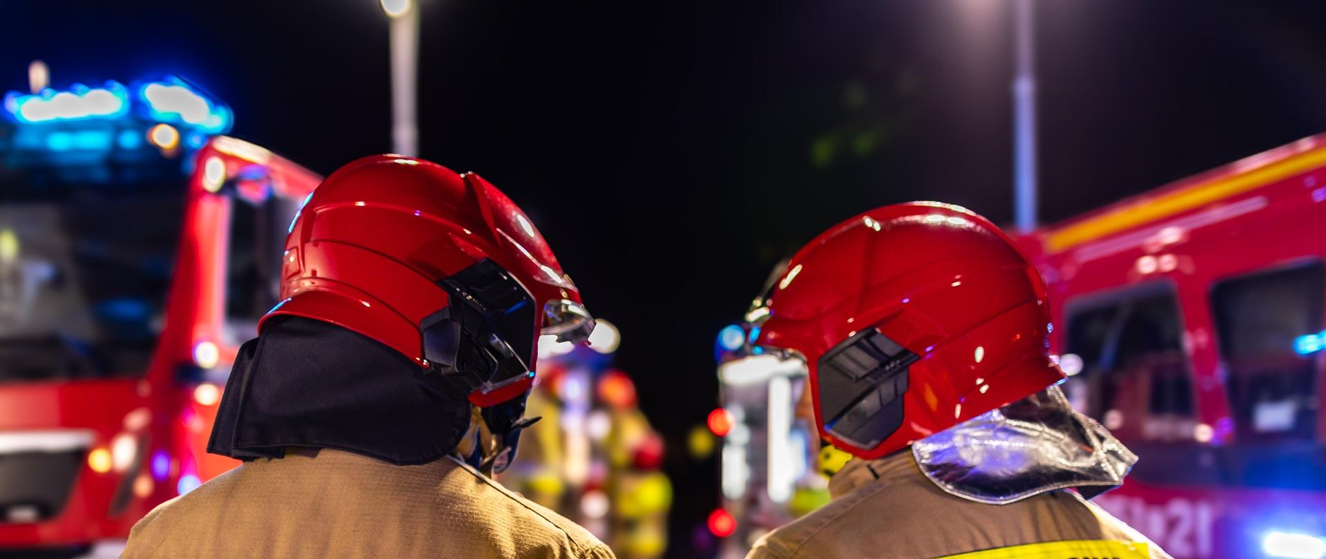 Na zdjęciu widzimy dwóch strażaków z Państwowej Straży Pożarnej (PSP) w pełnym umundurowaniu bojowym. Obaj są odwróceni tyłem do aparatu, a na plecach ich kurtek widnieje wyraźny napis: „PAŃSTWOWA STRAŻ POŻARNA”.