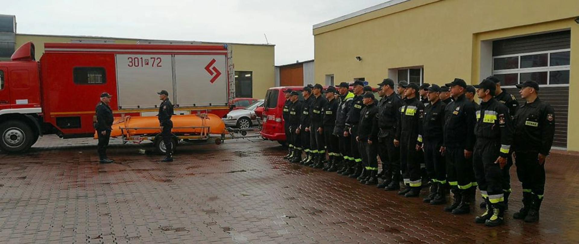 grupa strażaków stoi w dwuszeregu, obok czerwony samochód i pomarańczowy ponton, przy samochodzie na bacznosć stoi dwóch mężczyzn zwróconych do siebie twarzami