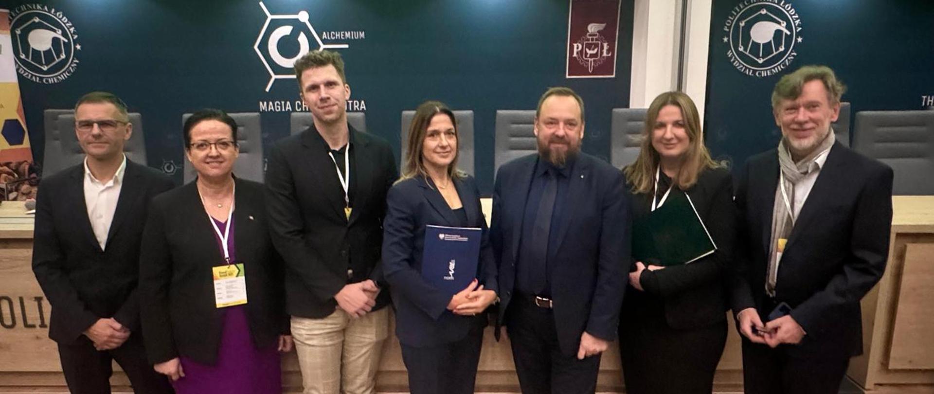 Na zdjęciu widać grupę siedmiu osób stojących razem w formalnym otoczeniu, prawdopodobnie podczas konferencji, spotkania naukowego lub oficjalnego wydarzenia.
Co dokładnie przedstawia zdjęcie:
Osoby są ubrane elegancko – w garnitury, marynarki i formalne stroje.
Dwie osoby trzymają w rękach teczki lub dokumenty, co sugeruje udział w prezentacjach, panelach lub podpisanie jakichś materiałów.
Wszyscy stoją przed stołem prezydialnym z mikrofonami i fotelami, co przypomina salę konferencyjną lub aulę wykładową.
W tle znajdują się logotypy wydziału chemicznego oraz nazwy związane z chemią („Alchemium”, „Magia chemiczna”), co wskazuje, że wydarzenie odbywa się najpewniej na uczelni technicznej.
Atmosfera jest oficjalna, a osoby wyglądają na uczestników lub organizatorów sesji naukowej, panelu lub ceremonii.