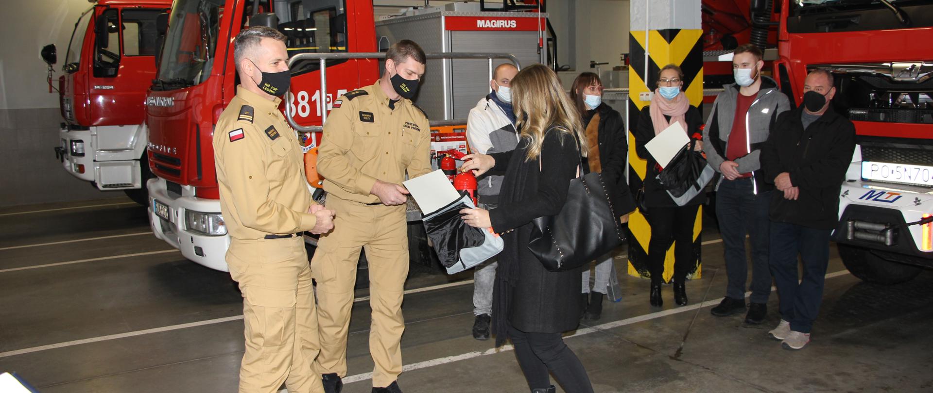 Strażacy wręczają nagrody osobą cywilnym stojącym w pomieszczeniu garażowym komendy. W tle pojazdy strażackie.