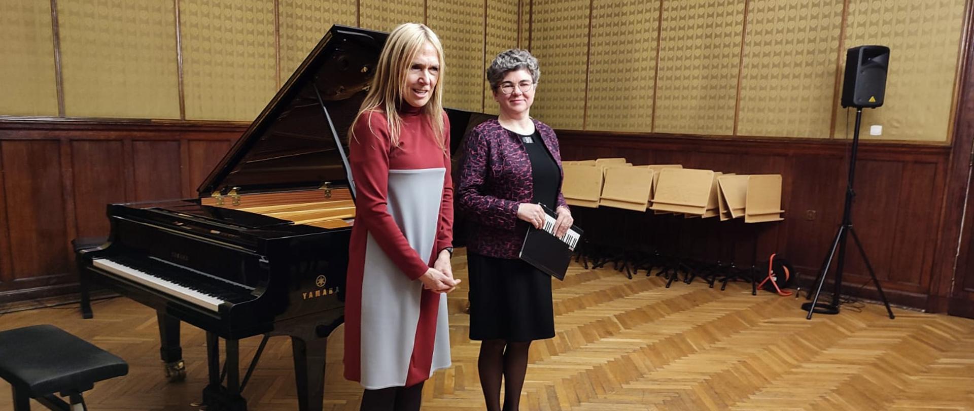 na zdjęciu dwie kobiety w kolorowych sukniach stojące przy czarnym fortepianie
