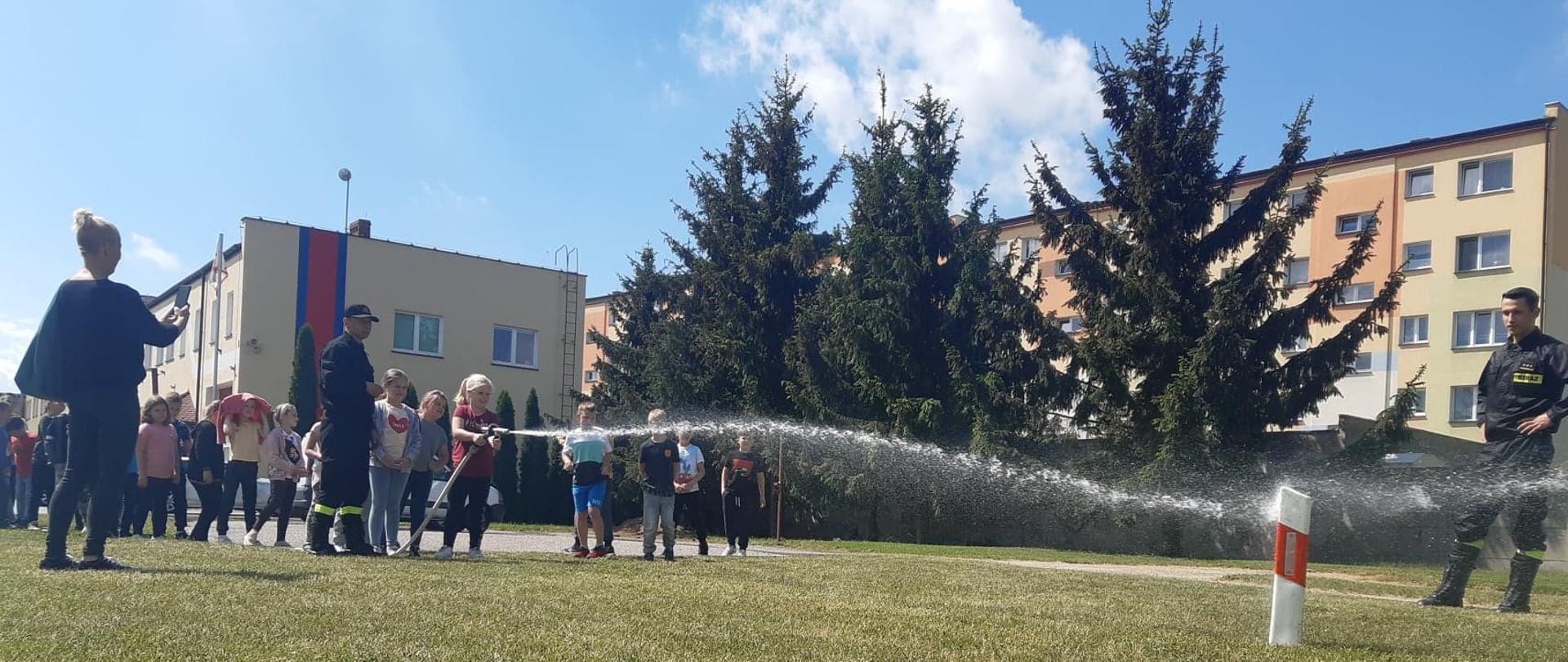 Dzieci z półkolonii organizowanych na terenie powiatu krotoszyńskiego odwiedziły strażaków. Zdjęcie wykonane na jednym ze stanowisk, na którym dzieci ustawione w kolejce korzystają z możliwości podania prądu wody za pomocą prądownicy. Całość asekuruje strażak. Opiekun dzieci wykonuje najmłodszym zdjęcia