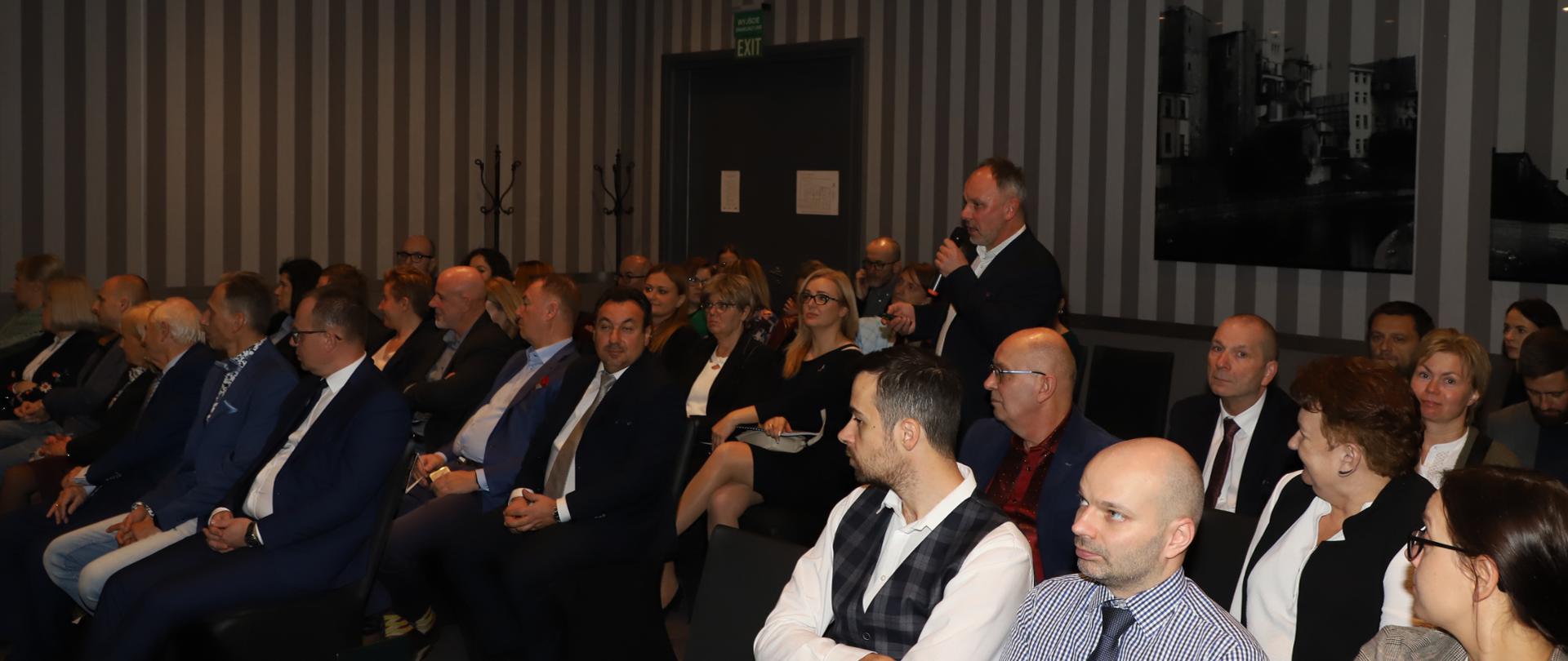 Mężczyzna stoi z mikrofonem a dookoła niego siedzi kilkadziesiąt osób