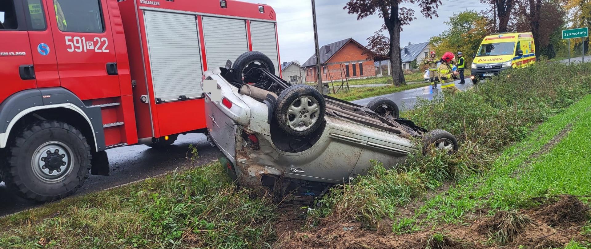 Srebrny samochód po dachowaniu jest w rowie, za nim samochód strażacki