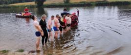 Strażacy w strojach kąpielowych oraz piankach ćwiczą łańcuch w zbiorniku wodnym, z tył widać dwie łodzie pontonowe.