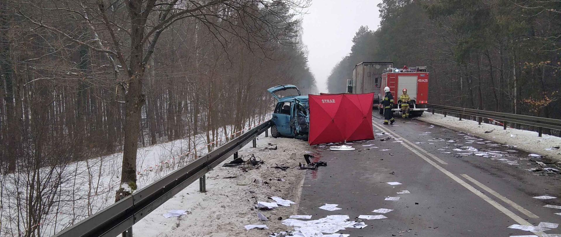Na zdjęciu droga asfaltowa z barierkami po obu stronach. Po prawej stronie samochód osobowy częściowo zastawiony czerwonym parawanem, nieco dalej po prawej stronie samochód strażacki a przed nim ciężarowy. Na jezdni porozrzucane kartki papieru. Po obu stronach drogi drzewa.