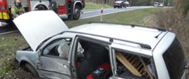 Srebrny samochód z otwartą pokrywą silnika, Urwane tylne lewe drzwi leżą obok. Wybite szyby.