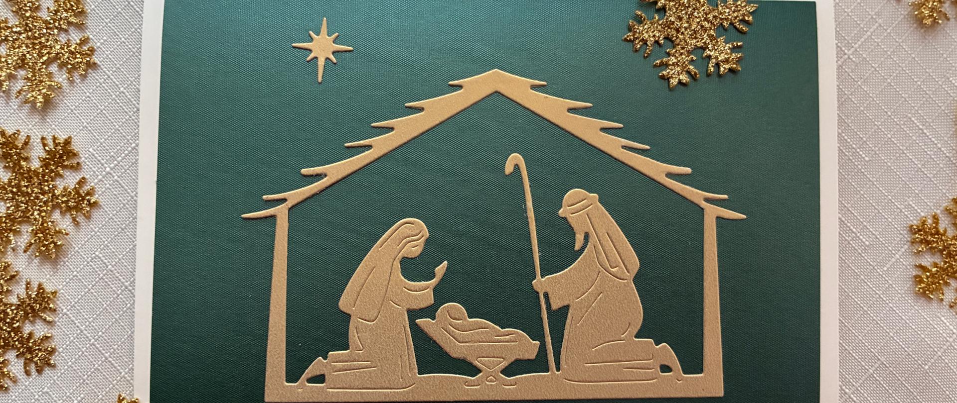 Karka świąteczna: Schemat szopki bożonarodzeniowej wycięty ze złotego papieru i naklejony na ciemnozielone tło. Kartka leży na jasnym obrusie wokół leżą złote płatki śniegu