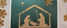 Karka świąteczna: Schemat szopki bożonarodzeniowej wycięty ze złotego papieru i naklejony na ciemnozielone tło. Kartka leży na jasnym obrusie wokół leżą złote płatki śniegu