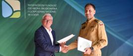 Dwóch mężczyzn, jeden w garniturze, drugi w mundurze strażackim, uściska sobie dłonie na tle baneru z napisem "WOJEWÓDZKI FUNDUSZ OCHRONY ŚRODOWISKA I GOSPODARKI WODNEJ W ŁODZI". Obydwaj trzymają dokumenty.