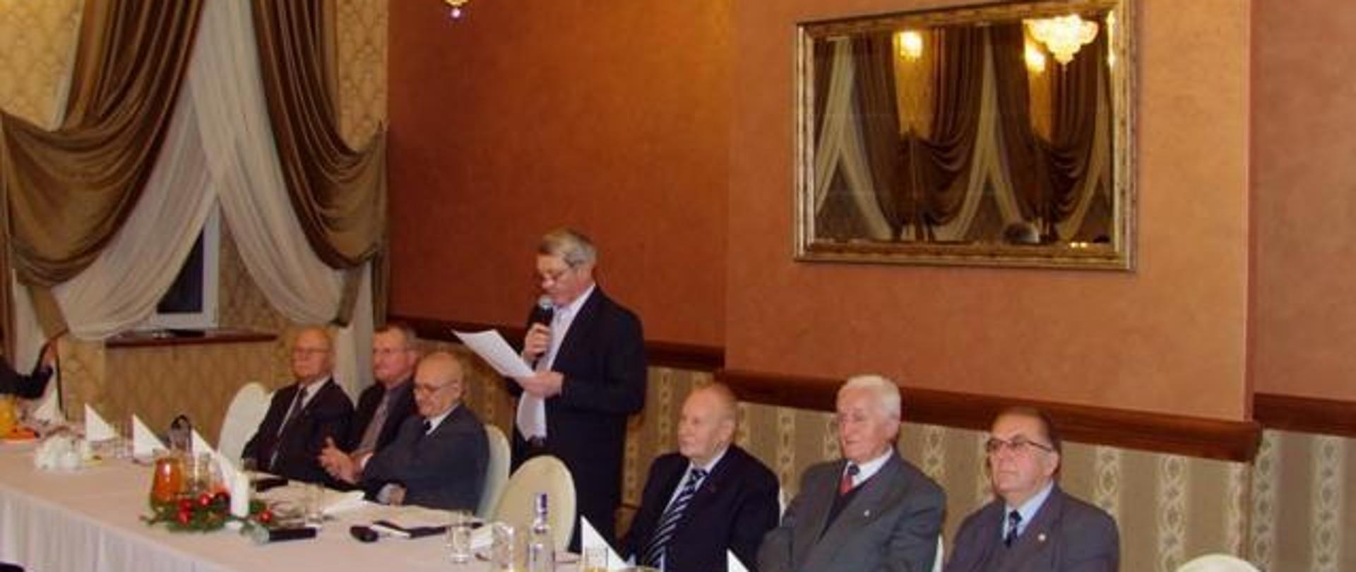 Na zdjęciu widzimy kilku mężczyzn siedzących przy stole prezydialnym. Mężczyzna po środku stoi i przemawia, w lewej dłoni trzyma białą kartkę natomiast w prawej czarny mikrofon zbliżony do ust. Mężczyźni w podeszłym wieku ubrani wyjściowo ciemne garnitury, jasne koszule widoczne są także krawaty. Stół nakryty białym obrusem, na stole widzimy także talerze, szklanki, serwetki, napoje. Dalej widzimy wystrój pomieszczenia w którym znajduje się spotkanie. Ściany wytapetowane na kolor pomarańczowy, na ścianie widzimy lustro w złotej oprawie.