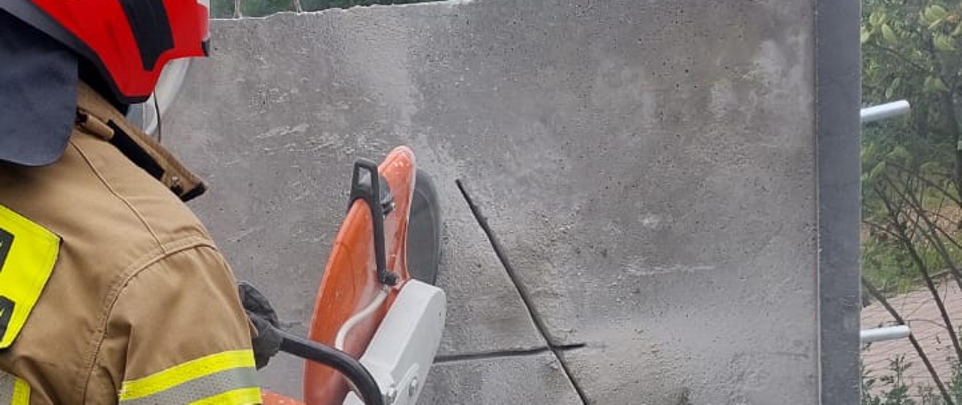 Strażak PSP w czerwonym hełmie używa przecinarki do cięcia grubego betonowego elementu. Z miejsca cięcia unosi się pył, a na powierzchni widoczne są wyznaczone linie. Ćwiczenie prezentuje techniki wykonywania dostępu w konstrukcjach betonowych podczas działań ratowniczych.