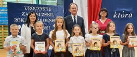 Siedmioro dzieci stoi w szeregu, trzymają w rekach książki, za nimi wiceminister Bernacki i dwie kobiety.