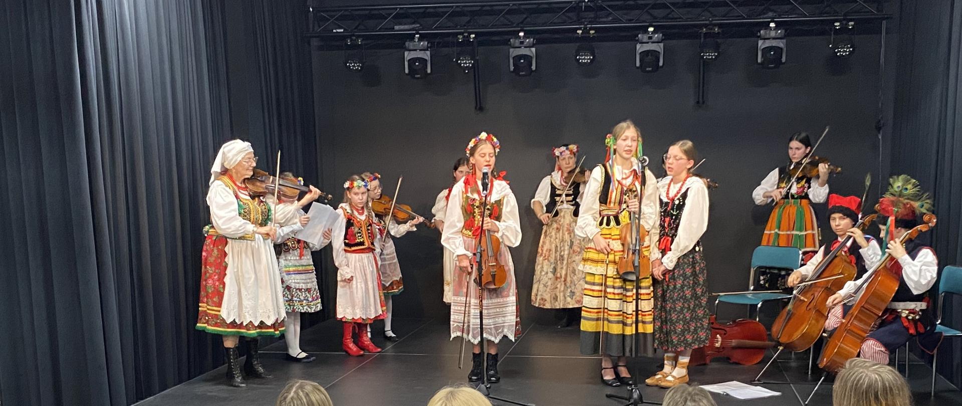 Na scenie w kręgu stoi kilkanaścioro dzieci i dwie kobiety, ubranych w kolorowe tradycyjne polskie stroje ludowe, grają na instrumentach smyczkowych. Po lewej stronie jest starsza kobieta w białej chustce na głowie. Po prawej stronie dwóch chłopców gra na wiolonczelach. Na środku stoją dwie dziewczyny i śpiewają do mikrofonów. Przed nimi widać od tyłu publiczność siedzącą na kolorowych fotelach.