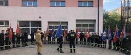 Widok na uczestników szkolenia i instruktorów, poczty sztandarowe OSP stoją w szeregu, przy ścianie budynku, przed nimi stoi dwóch instruktorów i jeden poczet sztandarowy
