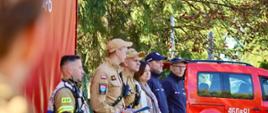 Grupa przedstawicieli służb ratunkowych i mundurowych stojąca w jednym rzędzie w plenerze. Po lewej stronie widać strażaka w mundurze bojowym z napisem „PSP”, obok niego dwóch strażaków w piaskowych mundurach służbowych i czapkach z daszkiem (jeden trzyma dokumenty), kobietę w stroju cywilnym z szalem oraz dwóch funkcjonariuszy policji w ciemnych mundurach i czapkach. W tle stoi czerwony samochód strażacki z numerem operacyjnym „460 P 91” na drzwiach oraz rosną zielone drzewa.