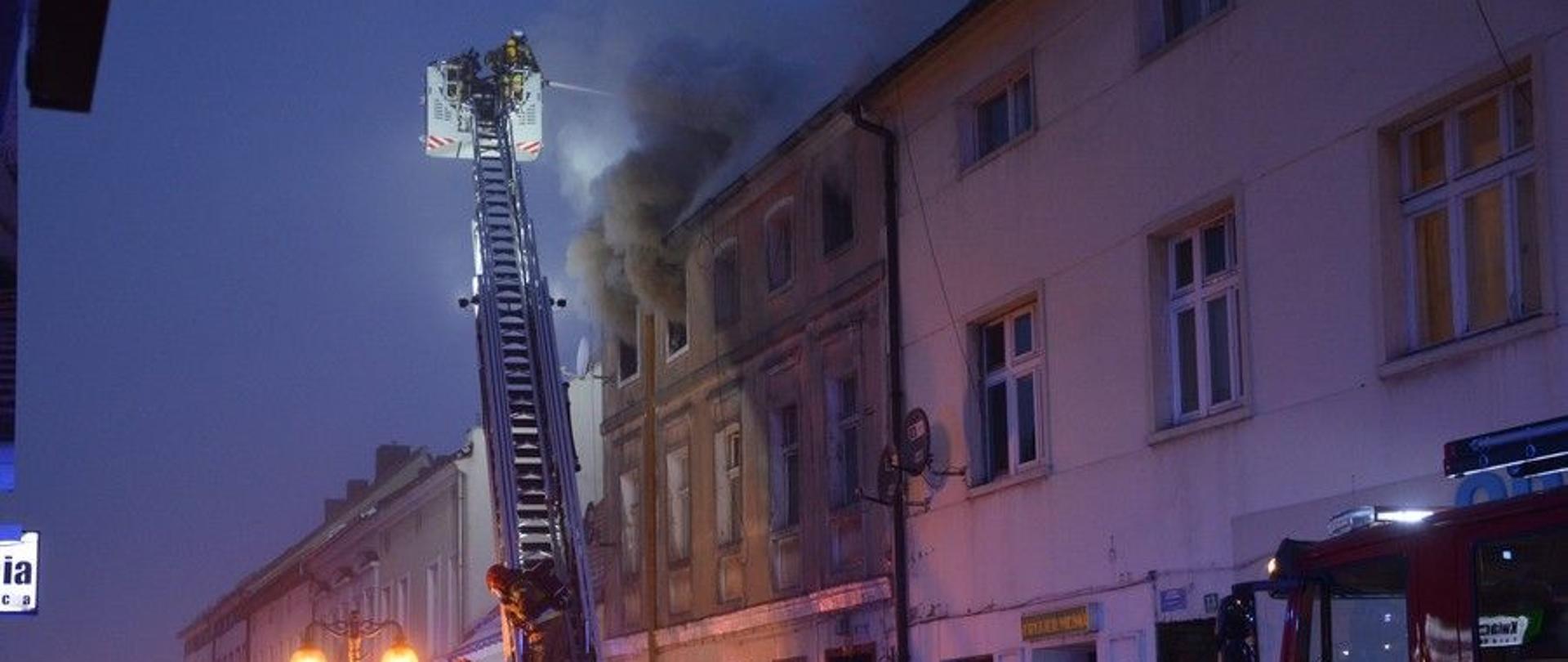 z okna kamienicy widobywa się dym przed budynkiem stoi wóz straży pożarnej i strazacy gaszący pożar