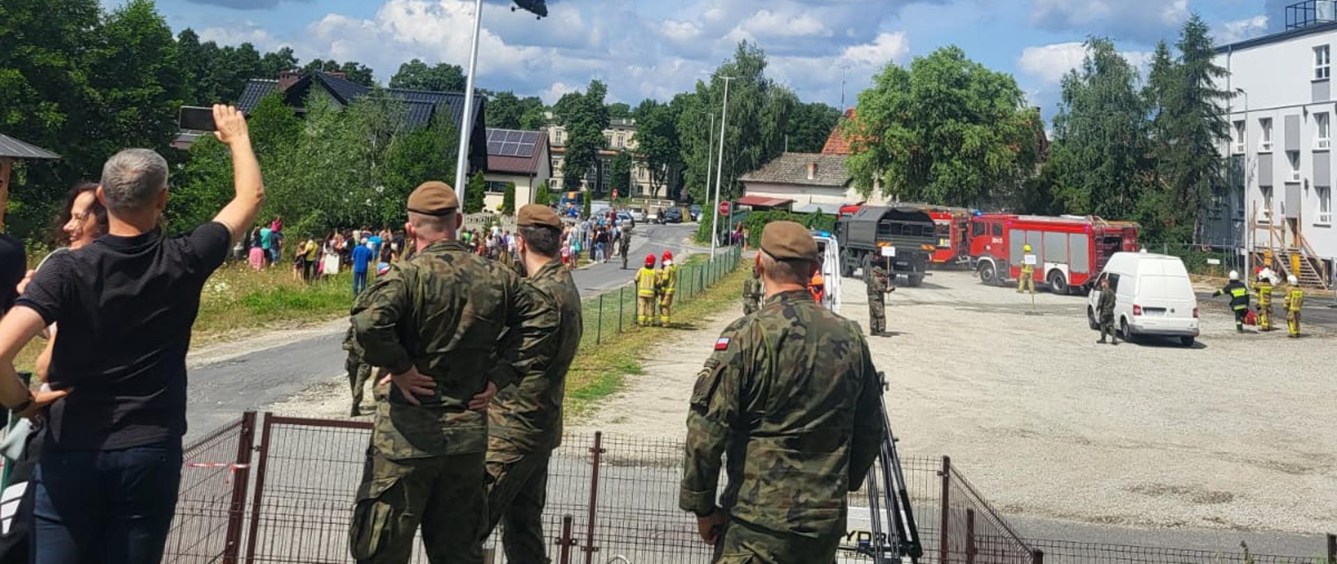 Strażacy, żołnierze i osoby cywilne stoją na placu i obok na ulicy.