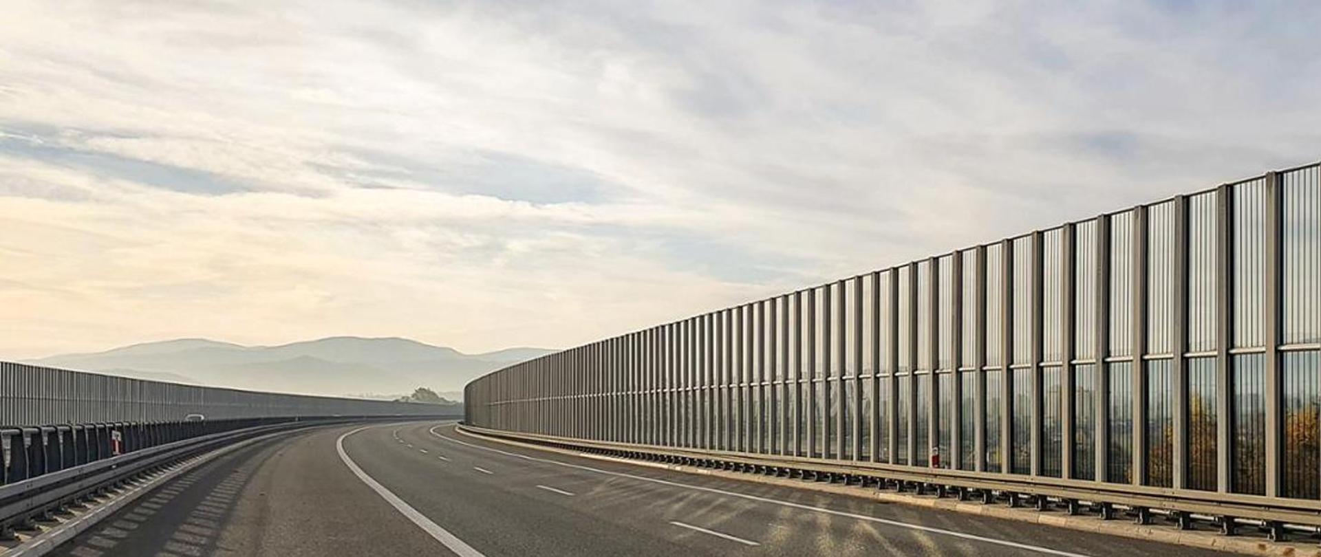 Widok na dwujezdniową drogę ekspresową z poziomu terenu. Droga przebiega w łuku poziomym. Na widnokręgu widoczne są góry, niebo jest zachmurzone. Drogę wyposażono w przezierne ekrany akustyczne oraz bariery ochronne.