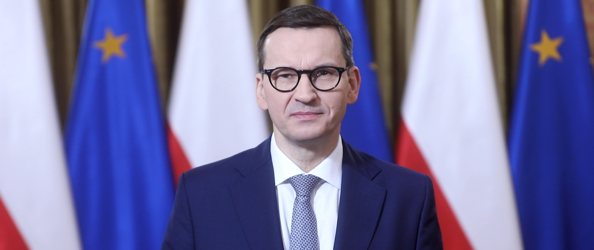 Prezes Rady Ministrów Mateusz Morawiecki na tle flag Polski i Unii Europejskiej