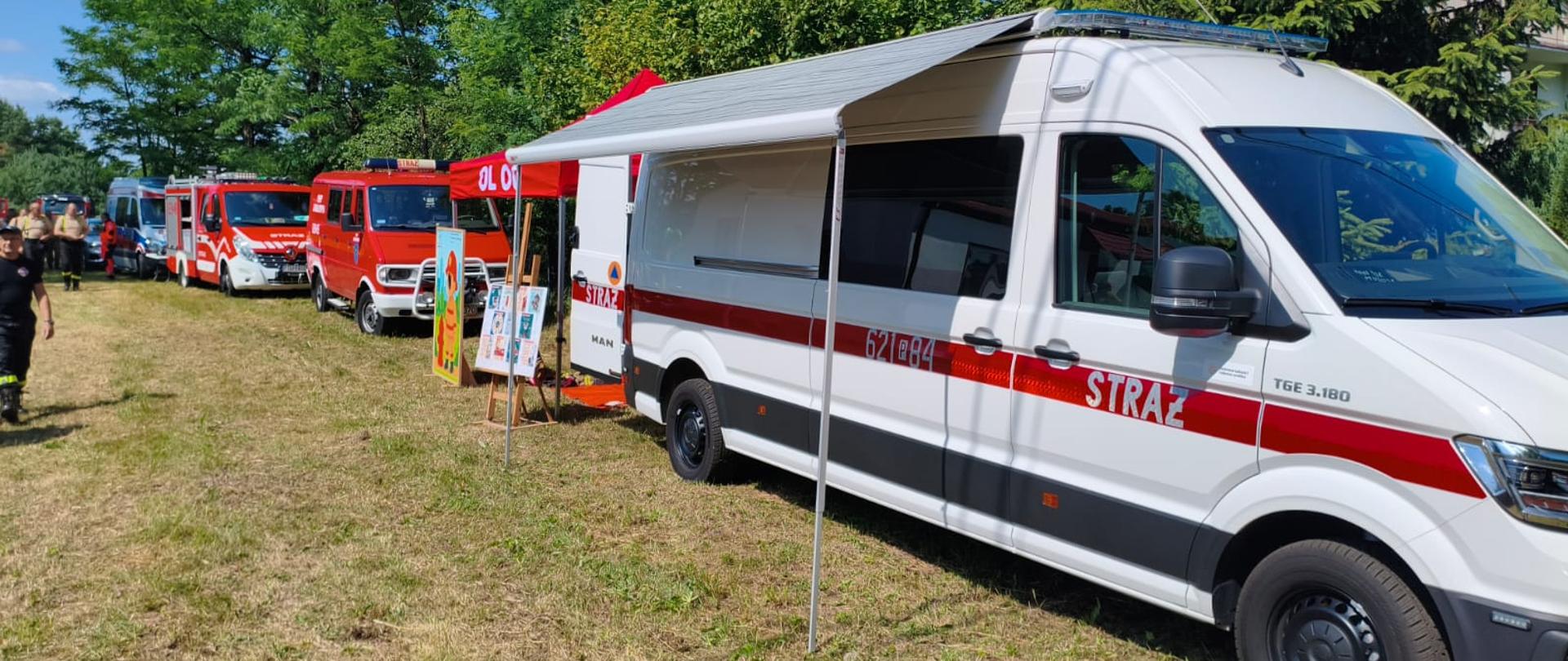 Na trawiastym terenie stoi kilka czerwonych i białych pojazdów straży pożarnej ustawionych w rzędzie. Na pierwszym planie widoczny jest biały samochód operacyjny z rozłożoną markizą, obok którego znajdują się tablice informacyjne i czerwony namiot. W tle widać strażaków oraz wysokie drzewa w słoneczny dzień. Pojazdy straży pożarnej prezentowane podczas plenerowego wydarzenia, z rozłożonym stoiskiem informacyjnym i namiotem na trawiastym terenie.