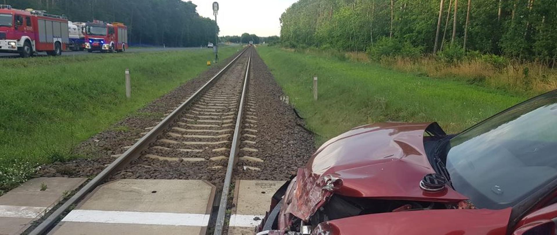 Na zdjęciu widać rozbity pojazd osobowy na przejeździe kolejowym oraz w oddali wozy strażackie 