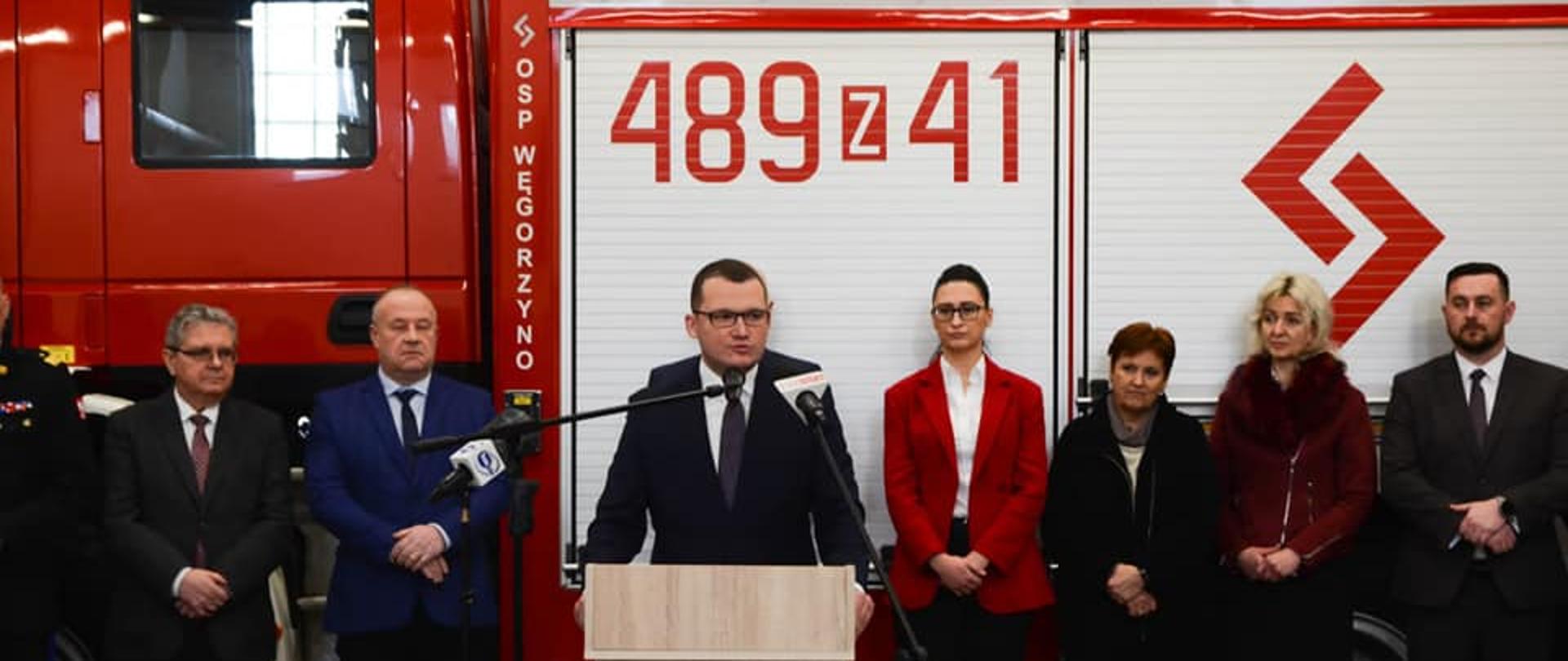 Przy mównicy sekretarz stanu w MSWiA Pan Paweł Szefernaker, za nim zaproszeni goście na tle samochodu ratowniczo gaśniczego.
