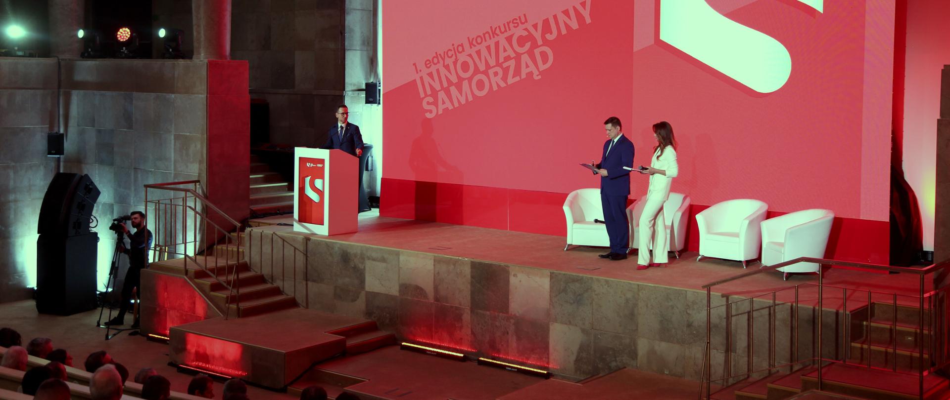 Na zdjęciu wiceminister Waldemar Buda stoi przy mównicy, po prawej stronie dwoje prowadzących, w tle ekran z napisem 1. edycja konkursu "Innowacyjny Samorząd".